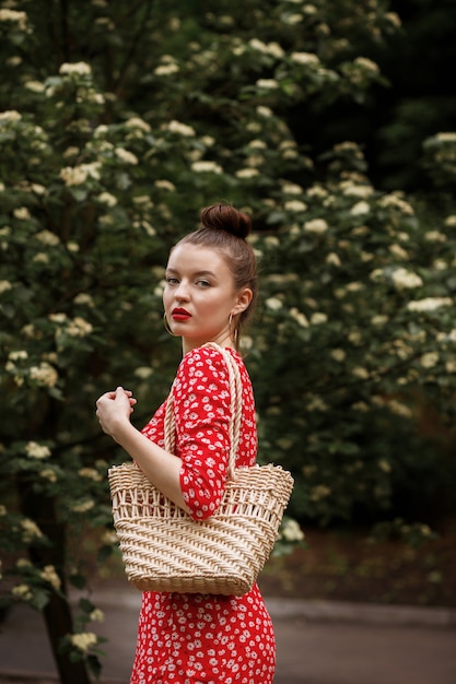 Mujer en un parque verde. vestido rojo de verano, una bolsa trenzada de paja con él. paseo de verano en el parque