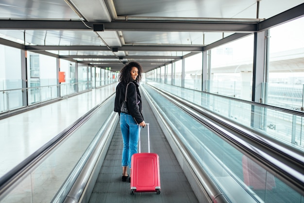 Mujer en la pasarela móvil en el aeropuerto con una maleta rosa.