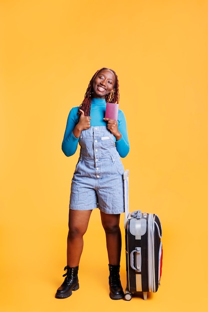 Mujer con pase de identificación mostrando como signo en la cámara, sintiéndose feliz de viajar en aventura. Modelo femenino que se va de viaje internacional de vacaciones con pasaporte y equipaje, pulgares hacia arriba.