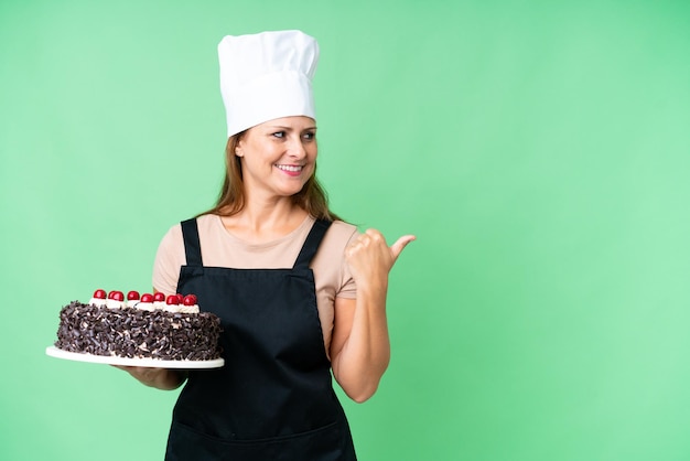 Mujer pastelera de mediana edad sosteniendo un gran pastel sobre un fondo aislado apuntando hacia un lado para presentar un producto