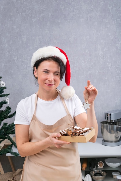 Foto mujer pastelera con sombrero de año nuevo y bata de cocina tiene caja de deliciosos dulces y guiños. marco vertical.