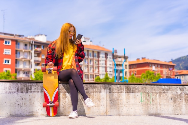 Mujer patinadora con una camiseta amarilla, camisa a cuadros roja y gafas de sol, sentada con patineta en la ciudad enviando un mensaje de audio con el teléfono