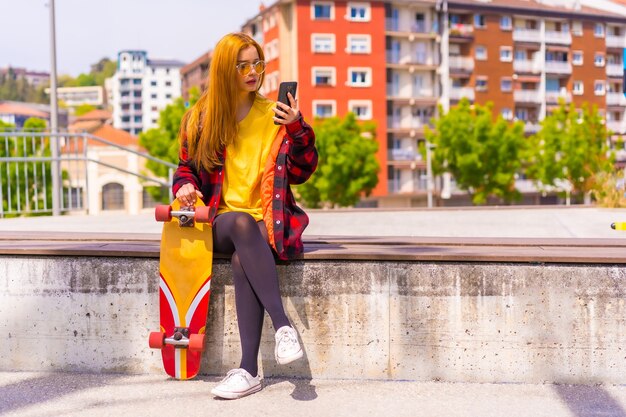 Mujer patinadora con una camiseta amarilla, camisa a cuadros roja y gafas de sol, sentada con patineta en la ciudad enviando un mensaje de audio con el teléfono