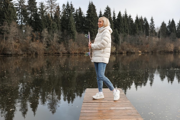Una mujer con una patineta camina en un parque de otoño junto al río aprendiendo a patinar pasando el fin de semana