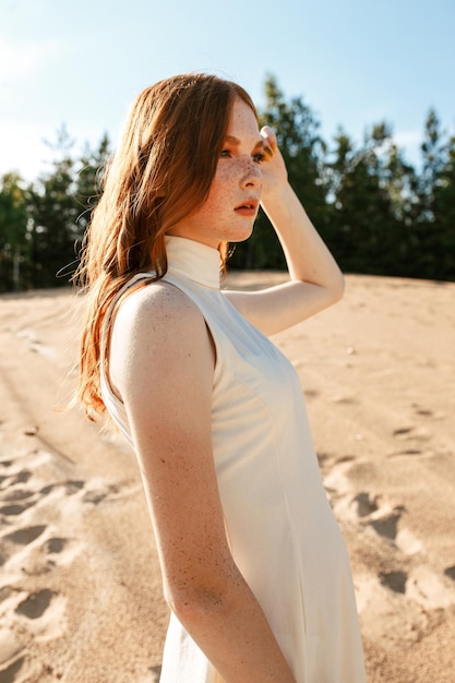 mujer con pecas y cabello largo y rojo de pie en un vestido blanco de verano en una colina arenosa