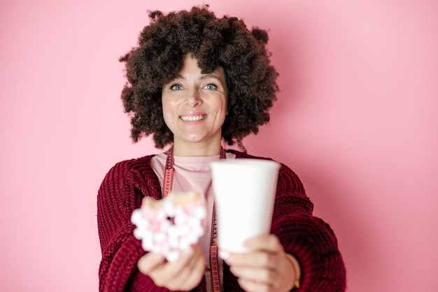 Mujer con peinado afro sostiene donut y bebida caliente de café o té en vaso de papel, adicción, cinta métrica alrededor del cuello, comida dulce, dieta poco saludable