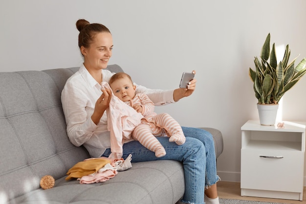 Mujer con peinado de moño con camisa blanca y jeans sentada tosiendo con su niño pequeño, revisa la ropa de bebé, sostiene el teléfono celular, graba videos o transmite en vivo.