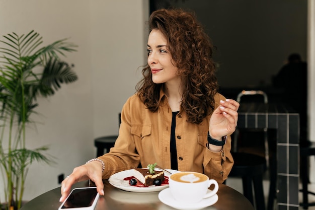 Mujer con peinado rizado vestida con blusa tierna está sentada con una taza de café y postre en el fondo de un elegante café moderno