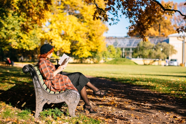 Mujer pelirroja en abrigo a cuadros y boina negra leyendo un libro en el banco descansando en el parque de otoño en sunn ...