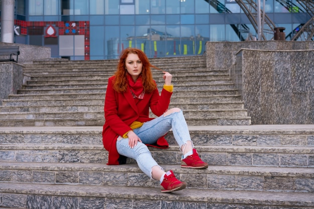 Una mujer pelirroja adulta joven con una chaqueta roja, jeans azules y zapatillas rojas está sentada en los escalones de ...