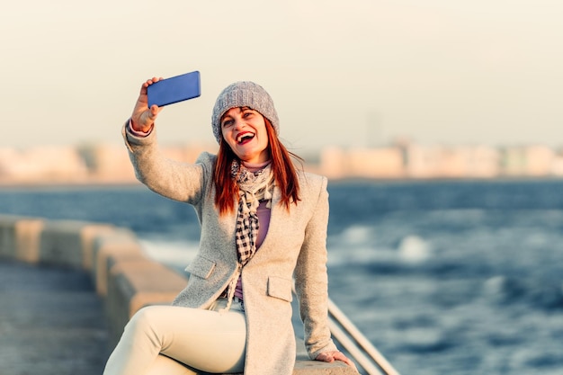 Mujer pelirroja al borde del mar se toma un selfie sonriendo a la cámara Gente