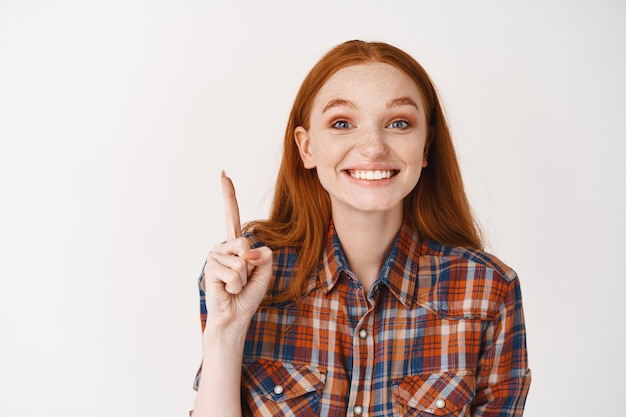 Mujer pelirroja alegre apuntando con el dedo hacia arriba y sonriendo con dientes blancos, mostrando publicidad, de pie sobre una pared blanca