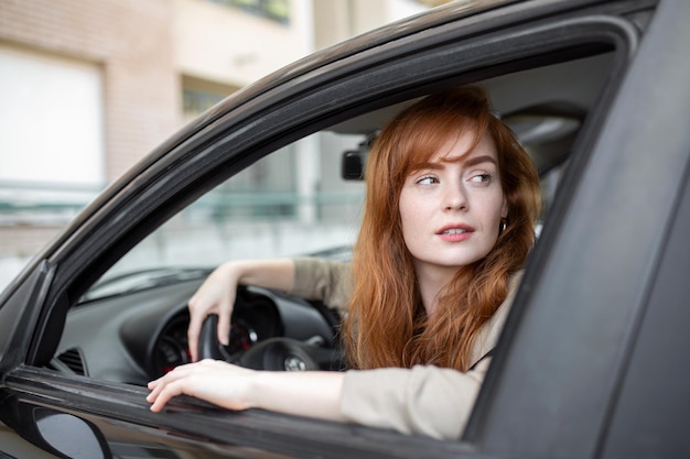 Mujer pelirroja alegre dentro del auto mirando hacia atrás desde el asiento del conductor mientras conduce durante el día