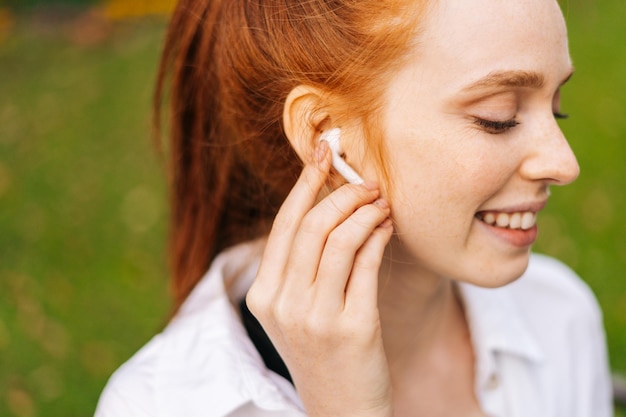 Mujer pelirroja alegre con los ojos cerrados escuchando música usando auriculares blancos inalámbricos al aire libre