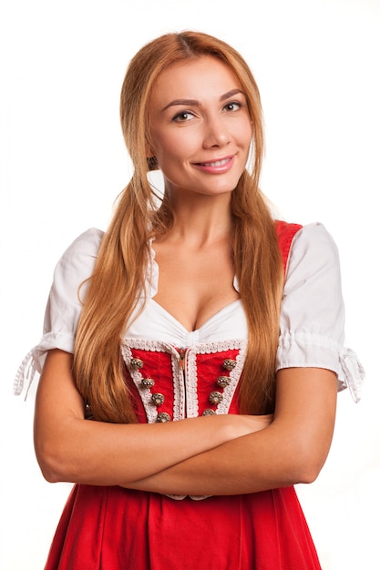 Mujer pelirroja atractiva magnífica en vestido bávaro tradicional que sonríe a la cámara con sus brazos cruzados aislados en blanco. Impresionante camarera de Oktoberfest