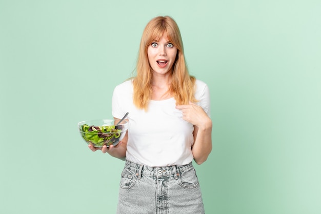 Mujer pelirroja bastante feliz y apuntando a sí mismo con un emocionado y sosteniendo una ensalada. concepto de dieta