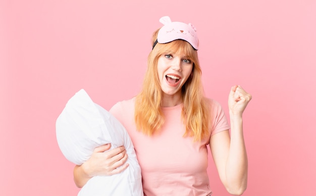 Mujer pelirroja bastante sorprendida, riendo y celebrando el éxito. en pijama y sosteniendo una almohada