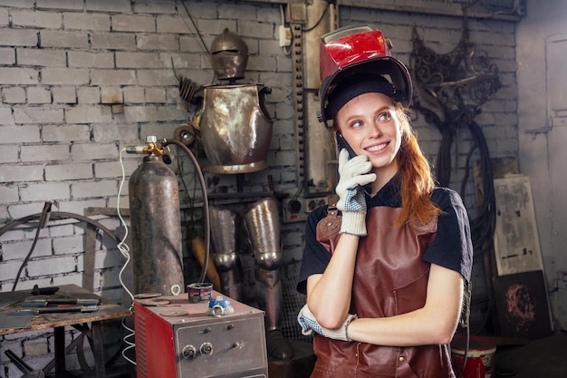 Mujer pelirroja bonita fuerte y digna con casco de protección y delantal de cuero con guantes que sostienen el concepto de pequeña empresa del instrumento de la máquina de soldadura