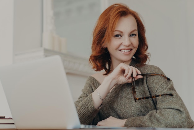 Mujer pelirroja calificada y exitosa sostiene anteojos para protección de la visión se sienta frente a una computadora portátil abierta posa en el espacio de coworking usa suéter usa un dispositivo moderno Concepto de trabajo a distancia