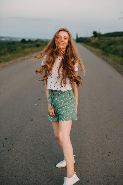 Mujer pelirroja caminando por un camino rural cerca del campo de trigo sonriendo a la cámara hermosa niña trigo fie...