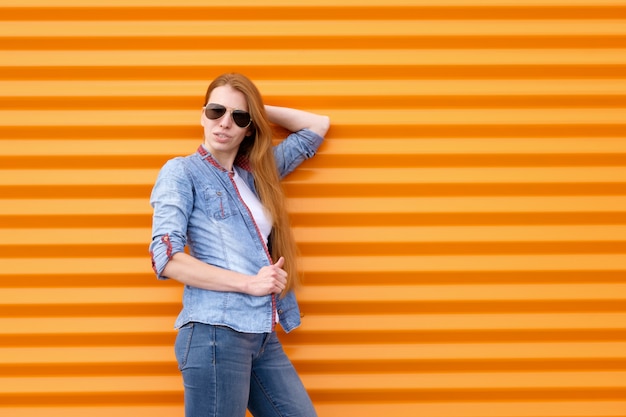 Mujer pelirroja en camisa de jeans con gafas de sol cerca de la pared naranja