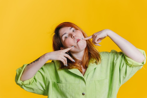 Una mujer pelirroja con una camisa verde se para sobre un fondo amarillo y hace muecas