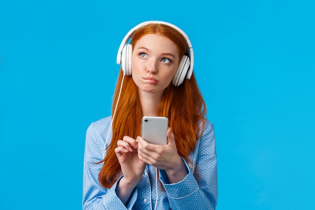 Mujer pelirroja caucásica pensativa y creativa, pensativa, chica astuta en ropa de dormir, mirando hacia arriba pensando, con auriculares escuchando música o podcast, sosteniendo el teléfono, pared azul
