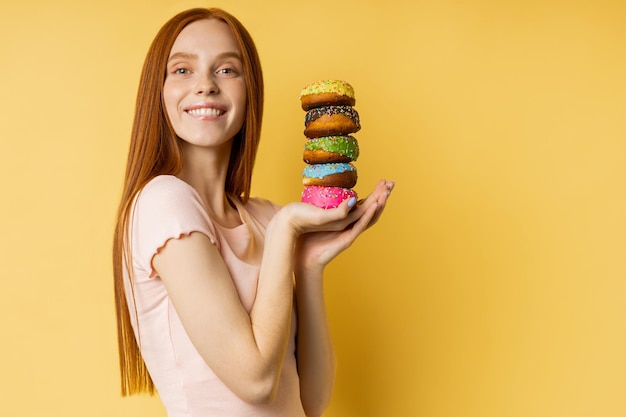 Mujer pelirroja complacida sosteniendo coloridos donuts decorados, sonriendo ampliamente mirando a cámara, disfrutando de dulces, fondo amarillo aislado con espacio de copia para su publicidad.