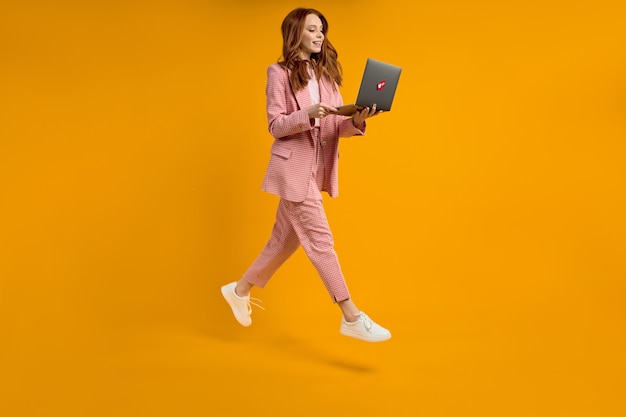 Mujer pelirroja correr salto escribiendo portátil vistiendo elegante traje rosa aislado sobre fondo amarillo en s ...