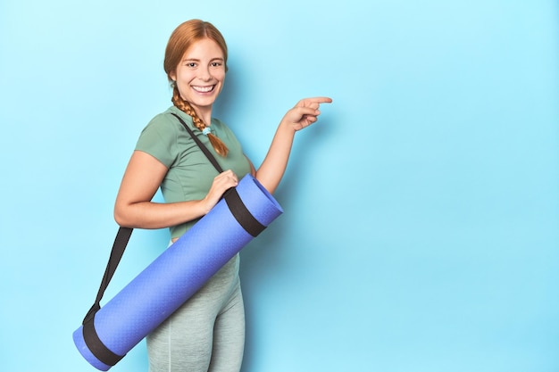 Foto mujer pelirroja deportiva llevando estera de yoga en el fondo azul del estudio