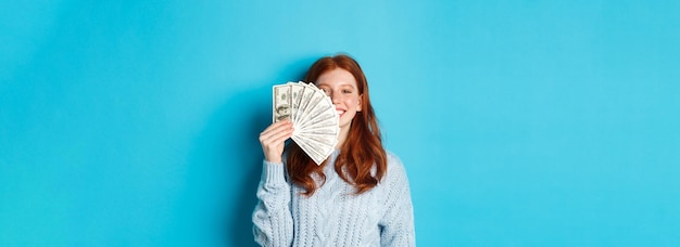 Foto mujer pelirroja encantada mostrando dinero cerca de la cara sonriendo satisfecha de pie lista para comprar sobre la persona