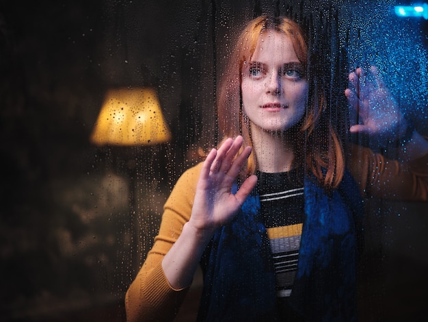 Mujer pelirroja se encuentra en la ventana con gotas de lluvia