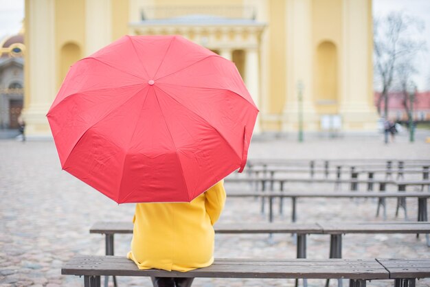 Una mujer pelirroja está sentada con la espalda en un abrigo amarillo con un paraguas rojo Una niña sentada sola en un banco en la catedral