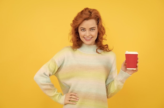 Mujer pelirroja feliz bebe café para llevar de la taza en la mañana de fondo amarillo
