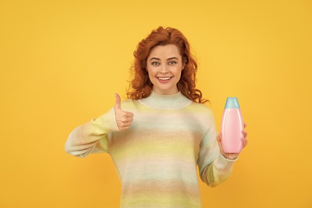 Mujer pelirroja feliz con cabello rizado sosteniendo champú o acondicionador mostrando el pulgar hacia arriba en la peluquería