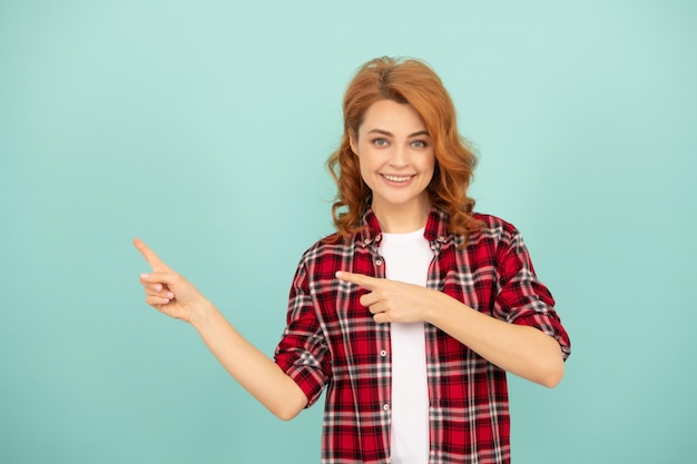 Mujer pelirroja feliz en camisa a cuadros señalar con el dedo sobre fondo azul.