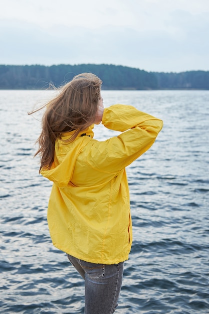 Mujer pelirroja en impermeable amarillo varado cerca del lago y mirando lejos. Nublado desagradable clima otoñal.