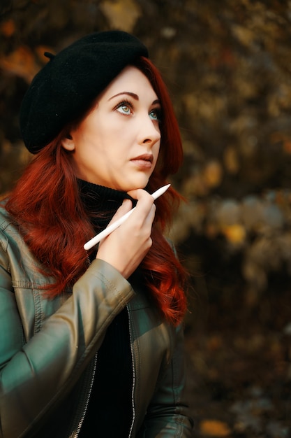 Mujer pelirroja inquietante en el parque mujer bonita en una boina y chaqueta de cuero hombre con un bolígrafo diseñado ...