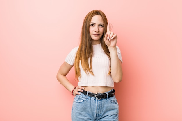 Mujer pelirroja jengibre joven contra una pared de color rosa que muestra el número uno con el dedo.