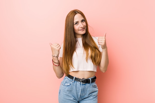 Mujer pelirroja jengibre joven contra una pared rosa levantando ambos pulgares, sonriente y confiado.