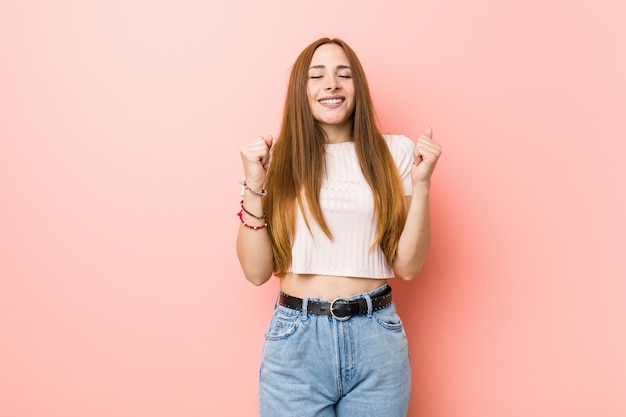 Mujer pelirroja jengibre joven contra una pared rosa levantando el puño, sintiéndose feliz y exitoso. Victoria