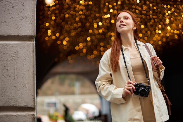 Mujer pelirroja joven muy de moda con paseo de la cámara en la ciudad, guirnaldas de boce en el fondo. Retrato de hermosa dama en chaqueta beige disfruta caminando sola al aire libre. Concepto de estilo de vida de personas