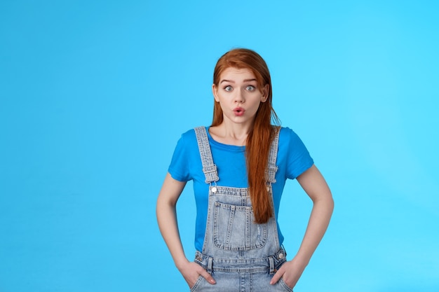 La mujer pelirroja linda nerviosa preocupada se siente ansiosa presionada antes de una entrevista importante, aguanta la respiración, exhala, hace pucheros sobre la situación difícil, parece insegura pararse con el fondo azul