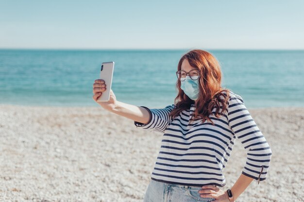 Mujer pelirroja con máscara con teléfono por la orilla del mar