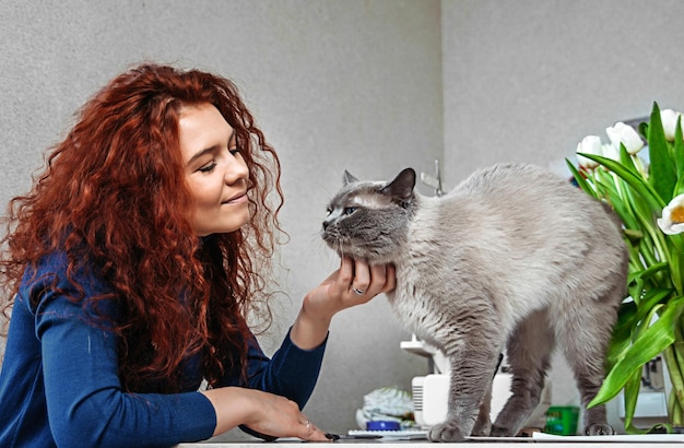 una mujer pelirroja de mediana edad rasca el cuello del gato, el gato impide que la anfitriona trabaje