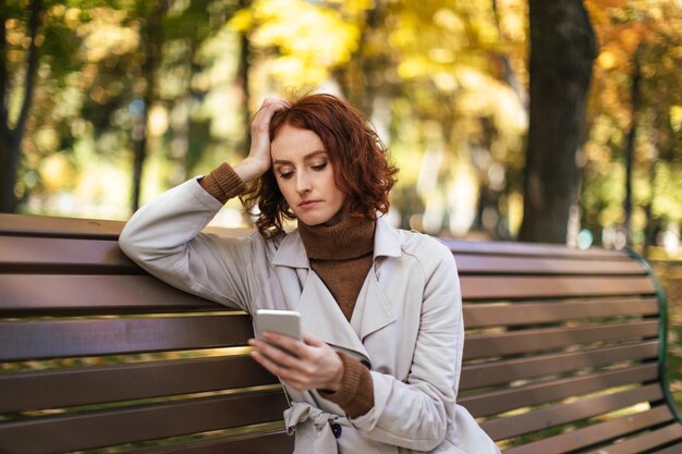 Mujer pelirroja milenaria caucásica infeliz en impermeable se sienta en el banco mira el teléfono inteligente en el parque