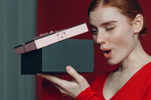 Mujer pelirroja mira a caja de regalo rosa con cinta Studio shot retrato fondo rojo