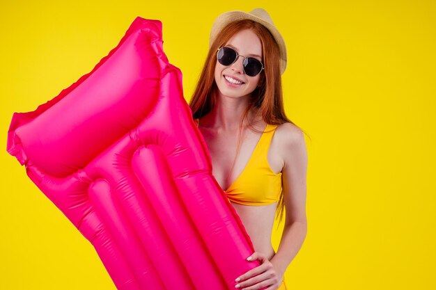 Mujer pelirroja pelirroja posando flotando con colchón inflable usando traje de baño y sombrero, guapa en el fondo amarillo del estudio.