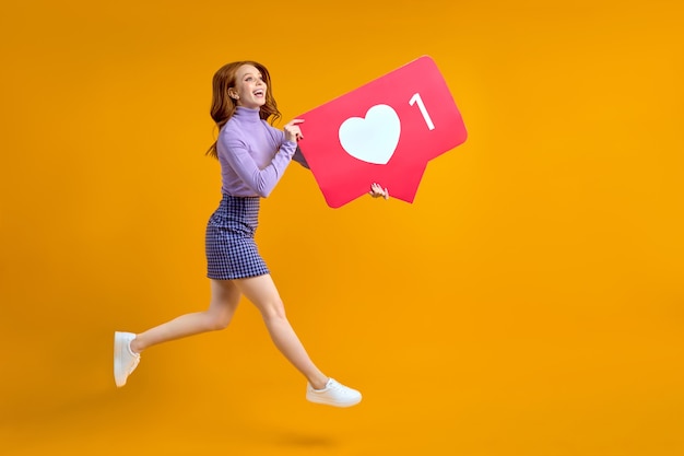 Mujer pelirroja persiguiendo gustos y apreciación saltando mujer manteniendo el icono del corazón de las redes sociales mirando copyspace aislado sobre fondo de color amarillo en estudio