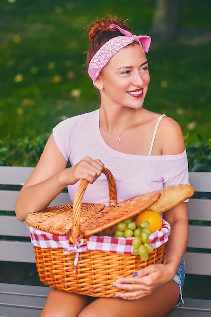 Mujer pelirroja positiva sentada en un banco con una cesta de picnic llena de frutas, pan y vino.